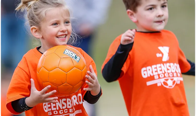 A young boy and girl wearing orange shirts that say Greensboro Soccer in white text. The girl is holding an orange soccer ball and smiling.